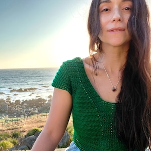 May include: A woman wearing a green crochet top with short sleeves and a silver necklace with a heart pendant. She is sitting on a wooden deck with a view of the ocean.