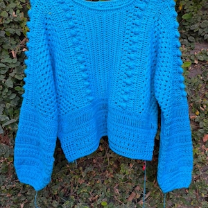 Puede incluir: Un suéter de manga larga en color azul brillante confeccionado con técnica de ganchillo. La prenda presenta un cuello redondo, filas verticales de puntos bobble en relieve y un patrón texturizado a lo largo del dobladillo y las mangas, en una percha amarilla.