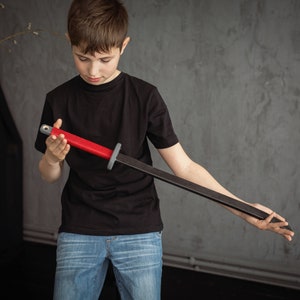 May include: A wooden toy sword with a red handle and a dark gray blade. The sword is held by a person wearing a black t-shirt and blue jeans. The background is a gray wall.