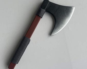 Pode incluir: Um pequeno machado com uma lâmina cinza escura e um cabo marrom. O cabo é enrolado com barbante cinza. A cabeça do machado é cinza escuro e a lâmina é prateada. Réplica em miniatura.
