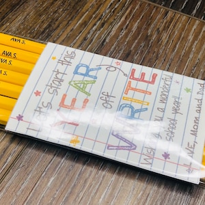 以下が含まれることがあります： ピンクの消しゴムと緑色のバンドが付いた黄色の鉛筆のパック。各鉛筆には「AVA S.」と書かれています。装飾カードには「Let's start this YEAR off WRITE. Wishing you a wonderful school year! LOVE, Mom and Dad.」と書かれています。