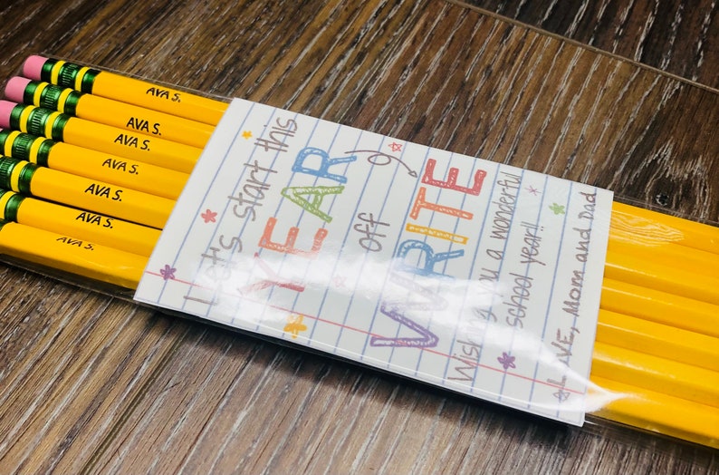 以下が含まれることがあります： ピンクの消しゴムと緑色のバンドが付いた黄色の鉛筆のパック。各鉛筆には「AVA S.」と書かれています。装飾カードには「Let's start this YEAR off WRITE. Wishing you a wonderful school year! LOVE, Mom and Dad.」と書かれています。
