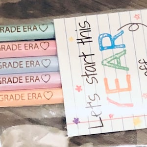 以下が含まれることがあります： 透明なビニール袋には、"IN MY SECOND GRADE ERA"の文字と小さなハートが付いた、パステルカラーの鉛筆が5本入っています。 "Let's start this YEAR off WRITE"という言葉が書かれたカードも含まれており、素晴らしい学校生活を願っています。