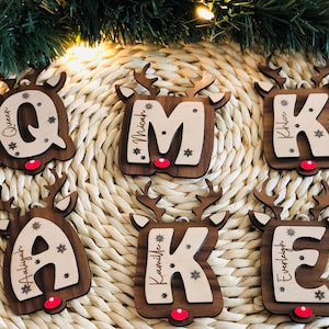 May include: Wooden Christmas ornaments shaped like reindeer, each featuring a letter and a name. The letters are in a light color, and the reindeer are brown with red noses. Snowflakes are also included in the design.