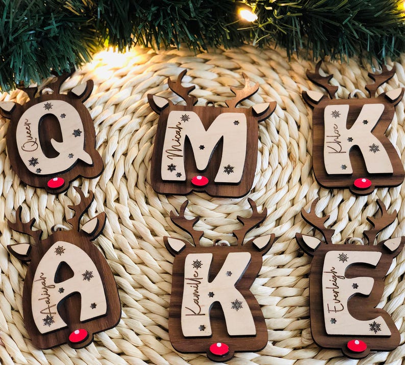 May include: Wooden Christmas ornaments shaped like reindeer, each featuring a letter and a name. The letters are in a light color, and the reindeer are brown with red noses. Snowflakes are also included in the design.