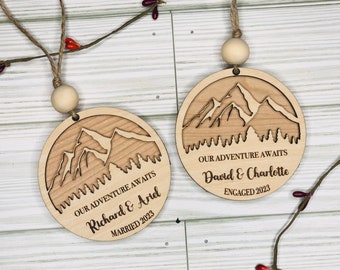 May include: Two wooden ornaments with a mountain range design and text that reads "Our Adventure Awaits". One ornament says "Richard & Ariel Married 2023" and the other says "David & Charlotte Engaged 2023".