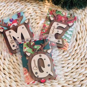 May include: Three wooden Christmas ornaments shaped like reindeer heads, each with a letter and a name. The letters are M, E, and Q. The names are Micah, Everleigh, and Queen. The ornaments are packaged in clear plastic and have festive backgrounds.