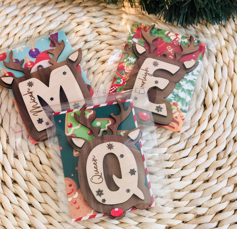 May include: Three wooden Christmas ornaments shaped like reindeer heads, each with a letter and a name. The letters are M, E, and Q. The names are Micah, Everleigh, and Queen. The ornaments are packaged in clear plastic and have festive backgrounds.