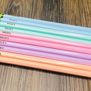 以下が含まれることがあります： 青、緑、ピンク、紫、ピーチなど、それぞれ異なる色合いのパステルカラーの鉛筆が10本。各鉛筆には「KHLOE E.」と刻印されています。鉛筆は木製の表面に配置されています。