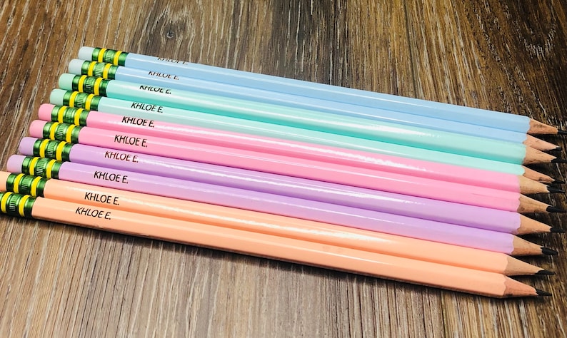以下が含まれることがあります： 青、緑、ピンク、紫、ピーチなど、それぞれ異なる色合いのパステルカラーの鉛筆が10本。各鉛筆には「KHLOE E.」と刻印されています。鉛筆は木製の表面に配置されています。