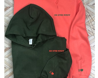 Puede incluir: Dos sudaderas con capucha, una en coral y otra en verde bosque, con el texto "XII-XVIII-MMXV" en cada una. La sudadera verde tiene un corazón rojo en la manga y la sudadera coral tiene un corazón verde en la manga.