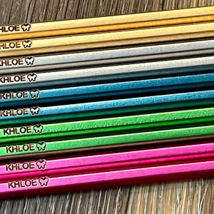 以下が含まれることがあります： 金、グレー、ティール、緑、ピンクなど、さまざまな色のパーソナライズされた鉛筆のコレクション。各鉛筆には「KHOLE」という名前と蝶のアイコンが刻印されています。鉛筆には緑と黄色のアクセントがあります。