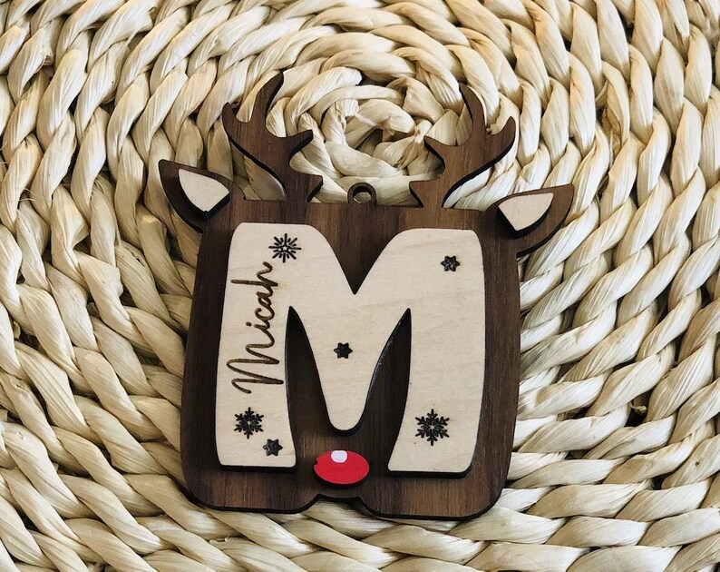 May include: Wooden Christmas ornament in the shape of a reindeer head. The ornament features a dark brown border, a light wood center with the letter "M", the name "Micah", snowflakes, and a red nose. The reindeer has antlers and ears.