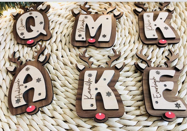 May include: Wooden Christmas ornaments in the shape of reindeer, each featuring a letter and a name. The letters are in a light wood color, and the reindeer are dark brown with red noses. Snowflakes are also included.