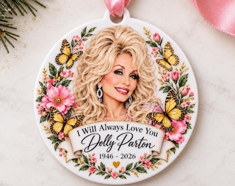 May include: A round white ceramic ornament featuring a portrait of Dolly Parton with blonde hair, surrounded by pink flowers and yellow butterflies. A banner at the bottom reads I Will Always Love You Dolly Parton 1946 - 2026 with a small gold heart.