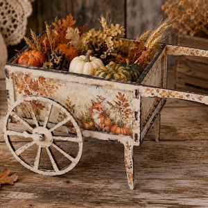 Op de afbeelding: Een decoratieve houten kar met een verweerde witte afwerking, voorzien van een bedrukte vos en herfstbladeren aan de zijkant. De kar is gevuld met kleine pompoenen, dennenappels en gedroogde tarwestengels als een seizoensgebonden decoratiestuk.