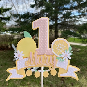 May include: A pink and yellow cake topper with a number one, lemons, and the name "Mary" on a white banner. The topper is on a white stick.