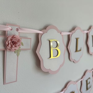 May include: A pink and white banner with the word "BLESS" in gold lettering. The banner includes a cross with a pink flower and a letter "B". The banner is strung with pink ribbon, suitable for a baptism or christening.