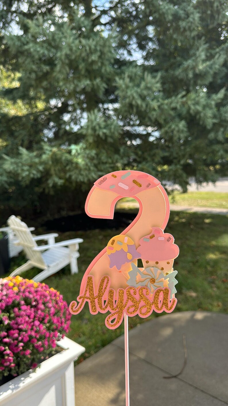 May include: A pink and gold number two cake topper with the name "Alyssa" in gold script. The number two is decorated with sprinkles and a cupcake design. The topper is on a thin pink stick.