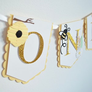 Peut inclure: Une bannière blanche et jaune avec le mot "ONE" en lettres dorées pailletées. La bannière est décorée d'abeilles, de fleurs et de nids d'abeilles.