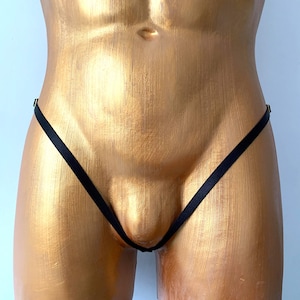 May include: A black thong featuring thin, minimalist elastic straps that form a V-shape across the front. The garment is displayed on a gold-toned torso mannequin, highlighting the simple construction and the dark fabric of the underwear.