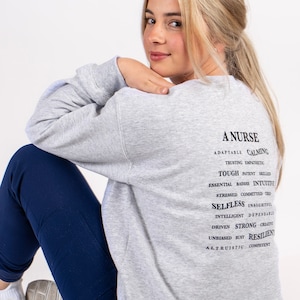 May include: A gray sweatshirt with the text "A Nurse" and a list of positive attributes associated with the profession. The text is in black and the sweatshirt is worn by a person sitting on the floor.