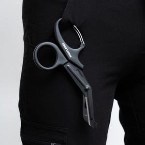 May include: Black medical shears with a curved handle and a silver-colored ring. The shears are partially inserted into a black pocket. The text "RNEXPLAINED" is printed on the handle.