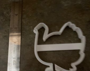 May include: A white plastic turkey-shaped cookie cutter next to a metal ruler. The ruler shows measurements in inches. The cookie cutter is approximately 3 inches wide and is designed with a horizontal bar across the body.