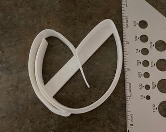 Może przedstawiać: Biała plastikowa wykrawaczka do ciastek w kształcie serca z poziomą linią. Obok wykrawaczki znajduje się linijka, pokazująca cale i centymetry. Wykrawaczka leży na ciemnej, teksturowanej powierzchni.