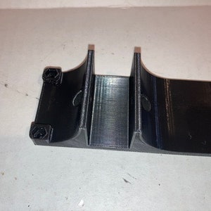 Op de afbeelding: Een zwart plastic 3D-geprint onderdeel met twee gebogen secties en meerdere zeshoekige gaten.