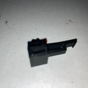 May include: Black plastic latch with a small lever. The latch has a rectangular shape and a small, raised lip on one side.