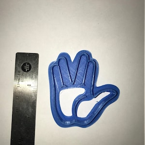 May include: A blue plastic cookie cutter shaped like a hand with four fingers raised in a Vulcan salute. The cookie cutter is about 2 inches wide.
