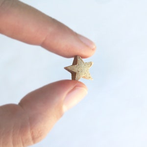 May include: A small, star-shaped object with a textured, light brown surface. The five-pointed star is held between two fingers against a white background. The object appears to be a decorative element or charm.