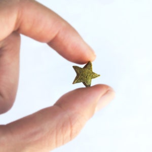 May include: A small, star-shaped object with a textured, olive-green appearance is held between two fingers against a white background. The star's edges are defined, and it appears to be made of a solid material.
