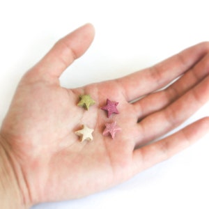 May include: Four small, star-shaped objects in shades of green, pink, and beige rest in an open palm against a white background. The stars have a textured appearance, suggesting they may be made of a natural material.