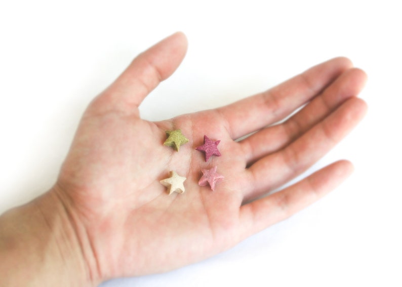 May include: Four small, star-shaped objects in shades of green, pink, and beige rest in an open palm against a white background. The stars have a textured appearance, suggesting they may be made of a natural material.