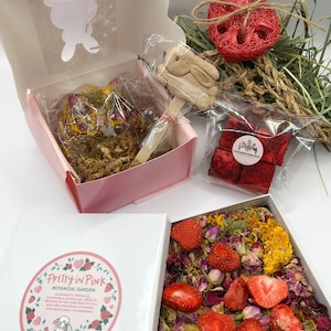 May include: A gift box set featuring a variety of botanical treats. Includes a pink box with a bunny cutout, a heart-shaped treat, a bunny-shaped cookie, and a box filled with dried strawberries and flowers. The set is labeled "Pretty in Pink Botanical Garden."