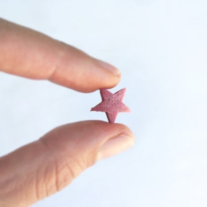 May include: A small, star-shaped object in a muted pink hue is held between two fingers against a white background. The star has five points and a slightly textured surface, suggesting it might be made of a natural material.