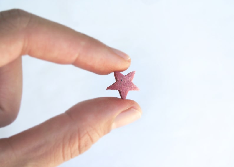 May include: A small, star-shaped object in a muted pink hue is held between two fingers against a white background. The star has five points and a slightly textured surface, suggesting it might be made of a natural material.