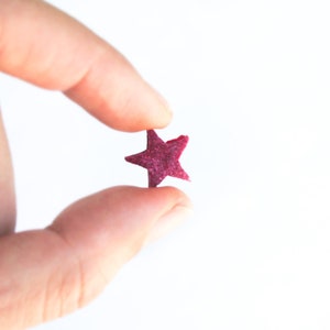 May include: A small, magenta-colored star held between fingers against a white background. The star has five points and a textured surface, appearing to be made of a soft material. The image is well-lit, highlighting the star's color and shape.