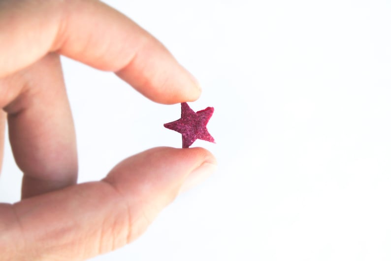 May include: A small, magenta-colored star held between fingers against a white background. The star has five points and a textured surface, appearing to be made of a soft material. The image is well-lit, highlighting the star's color and shape.