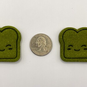 May include: Two green felt toast-shaped ornaments with a smiling face. The ornaments are about the size of a US quarter.