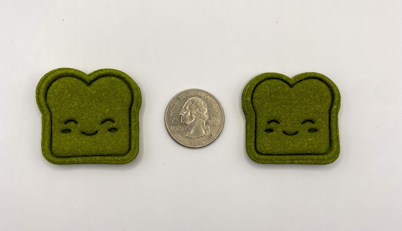 May include: Two green felt toast-shaped ornaments with a smiling face. The ornaments are about the size of a US quarter.