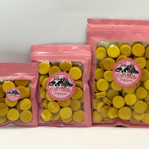 Op de afbeelding: Drie roze hersluitbare zakjes gevuld met gele ronde lekkernijen. De zakjes hebben een wit label met de tekst "Fluffybun Confections" en een cartoonkoe met een roze strik.