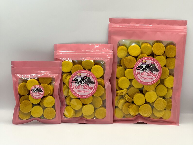Op de afbeelding: Drie roze hersluitbare zakjes gevuld met gele ronde lekkernijen. De zakjes hebben een wit label met de tekst "Fluffybun Confections" en een cartoonkoe met een roze strik.