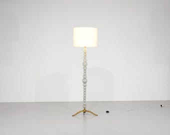May include: A modern floor lamp with a white lampshade and a beaded, light gray stem. The lamp has a gold-colored, three-legged base and a power cord. The lamp is a decorative lighting fixture.