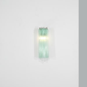May include: A wall-mounted light fixture with a vertical design. The fixture is composed of multiple, parallel, light green glass rods. A bright light shines through the center of the fixture, illuminating the surrounding area. The fixture is mounted on a white wall.
