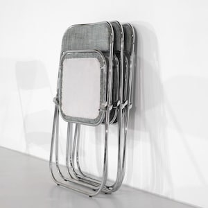 May include: A stack of three folding chairs with chrome frames and gray fabric seats and backs. The chairs are leaning against a white wall, showcasing their design and construction. The seats have a white center.
