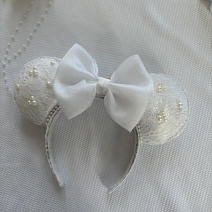 May include: White mouse ear headband featuring a satin bow and lace-covered ears. The ears are decorated with small pearls, and the headband is silver-trimmed. A decorative accessory.