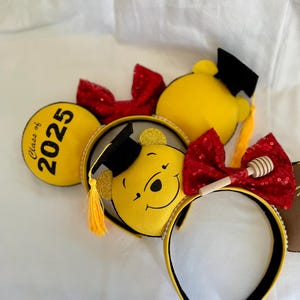May include: Yellow Minnie Mouse ears with a Winnie the Pooh graduation cap and a red bow with a honey pot charm. The left ear has the text "Class of 2025" on it.